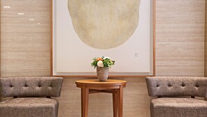 A lobby with two upholstered armchairs, a wooden table with a vase of flowers, and a large abstract painting on the wall.