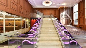 A hotel interior with a central staircase flanked by glass railings and purple lighting, wooden paneling on the walls, and a reception area with a sign reading "Hotel Skypark Central".