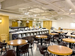 A spacious dining area with multiple round tables and chairs, a buffet setup with various food items, and a modern ceiling with exposed pipes and lighting fixtures.
