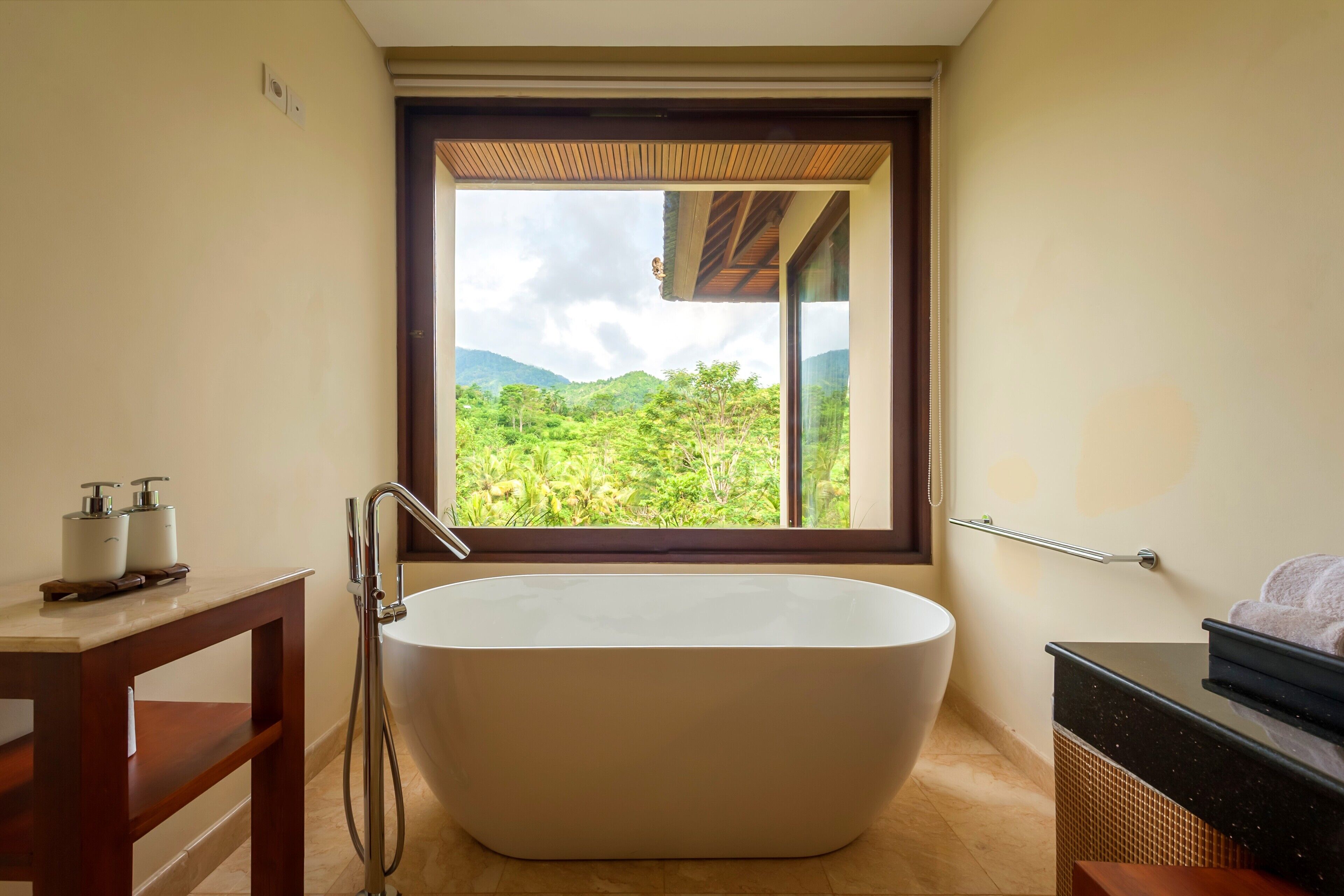 A modern bathroom with a freestanding bathtub, a wooden vanity, and a large window offering a view of lush greenery and mountains.