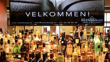 A bar area with a variety of liquor bottles and a television screen displaying 'VELKOMMEN!' with the Reenskaug Hotel & Restaurant logo.