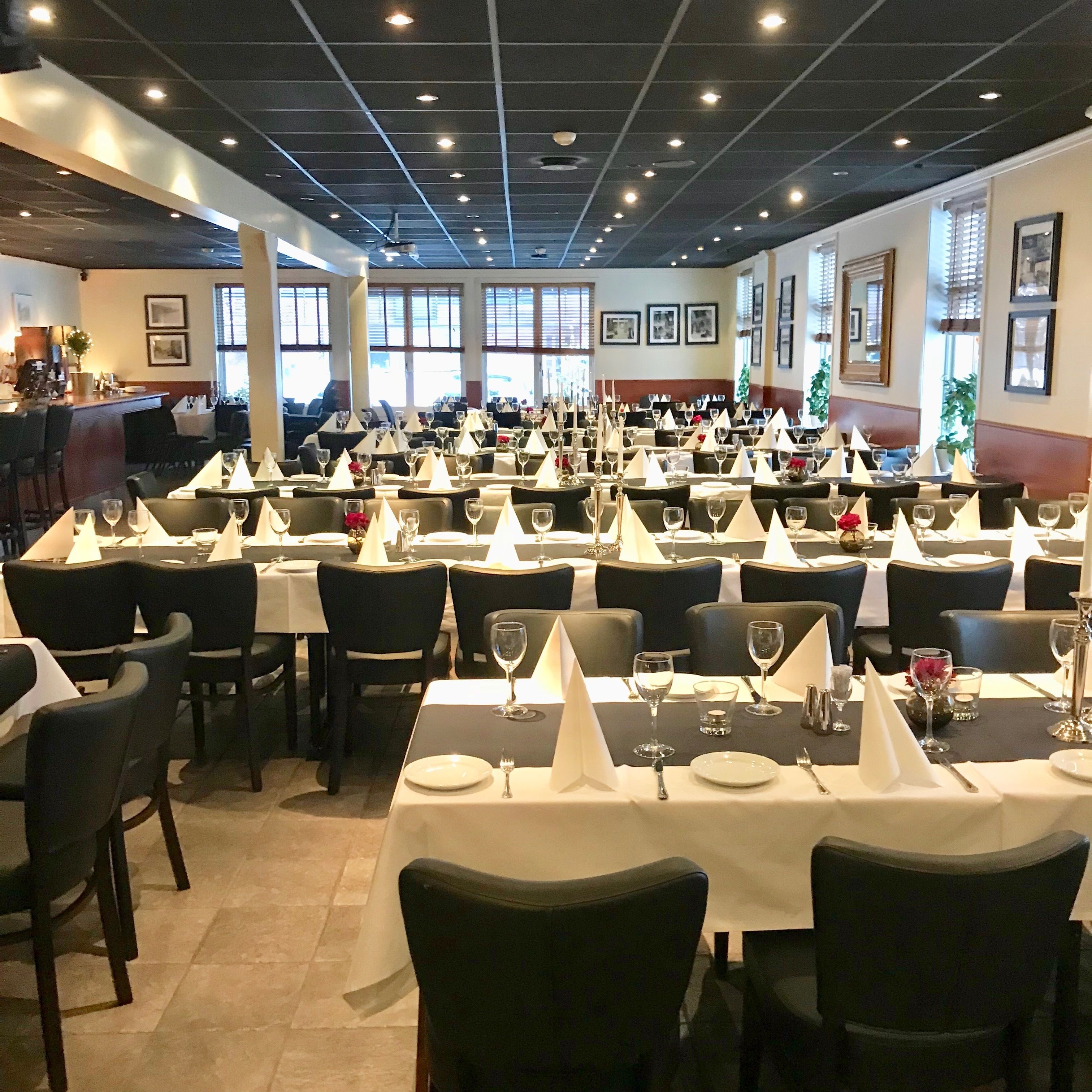 A spacious dining area with multiple tables set for service, featuring white tablecloths, neatly arranged chairs, and a bar area visible in the background.
