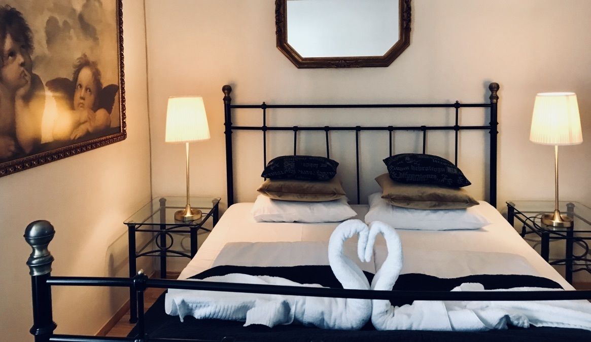 Una cama bien hecha con una toalla dispuesta en forma de corazón, dos lámparas de noche y una obra de arte enmarcada en la pared.