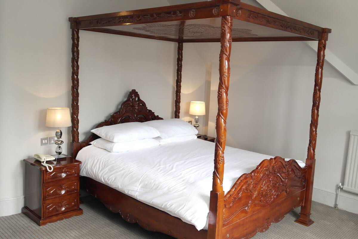 Una cama con dosel de madera con detalles tallados, flanqueada por mesitas de noche con lámparas.