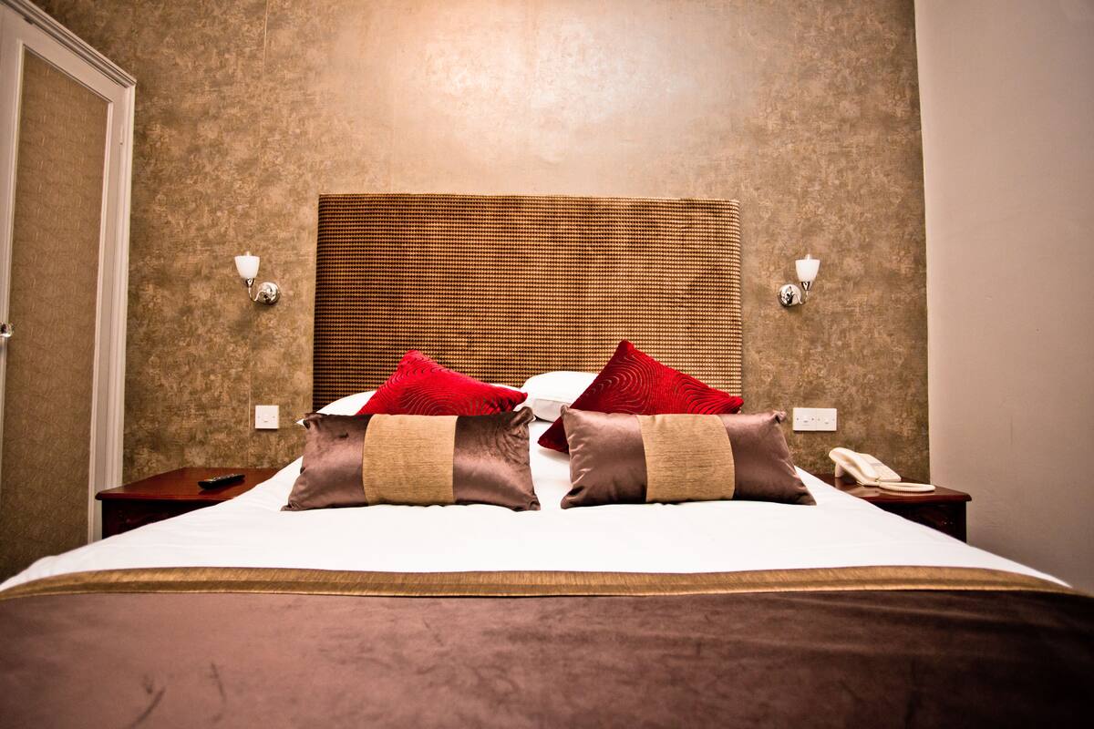 Una cama bien tendida con cabecera, dos mesitas de noche y un teléfono en la pared.