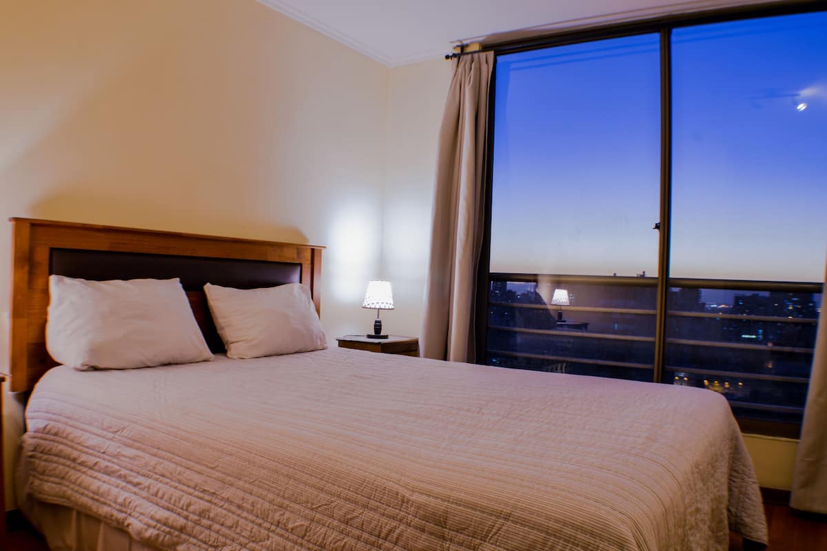 Ein Hotelzimmer mit einem großen Bett, einem Nachttisch mit Lampe und Blick auf die Stadt im Abendlicht durch ein großes Fenster.