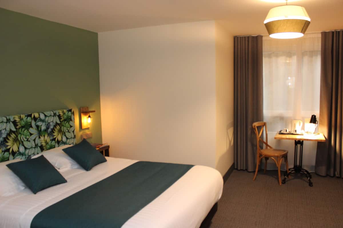A hotel room with a bed, a desk, a chair, a lamp, and a window with curtains.