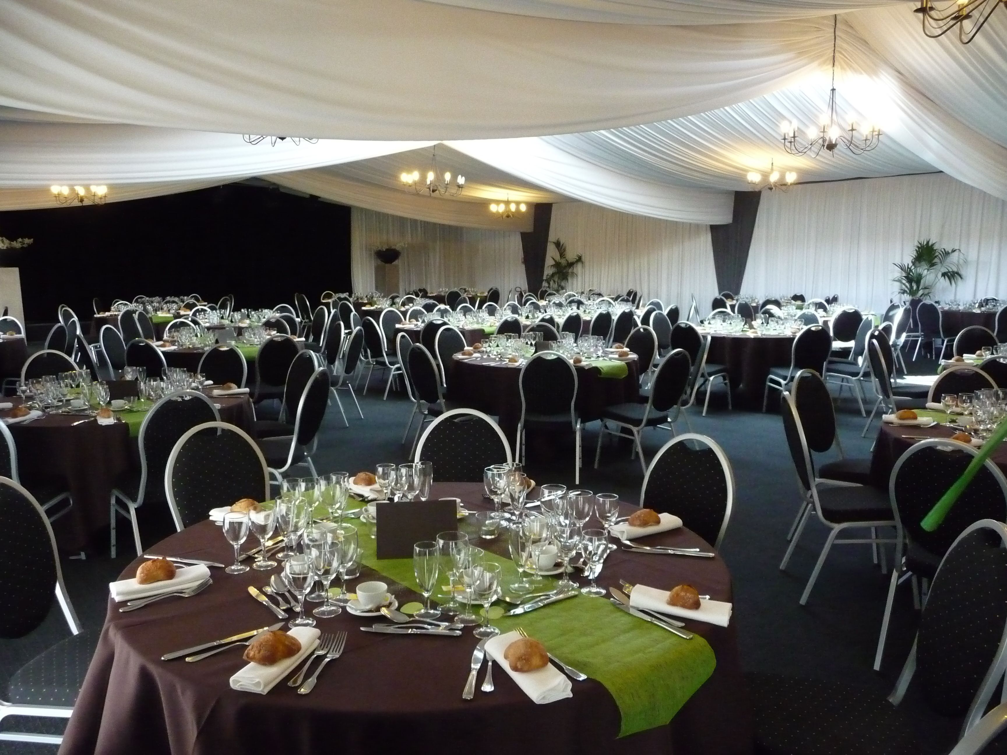 Un salón de banquetes con mesas redondas dispuestas para un evento formal, con manteles blancos, sillas negras y caminos de mesa verdes.