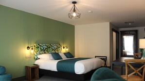 Een hotelkamer met een bed, een bureau en een stoel. Er staat een plant aan de muur en er hangt een plafondlamp.