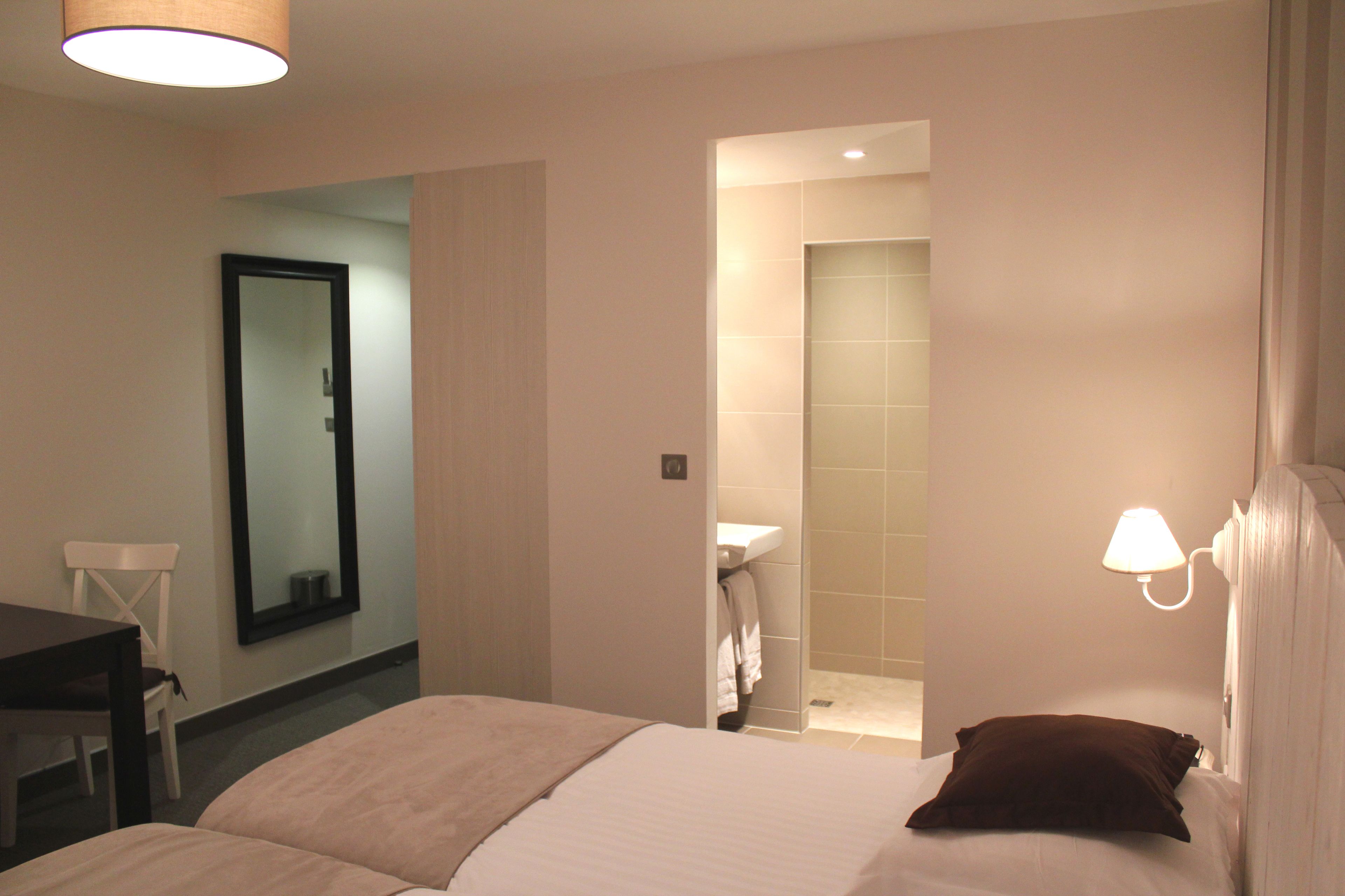 Une chambre d’hôtel avec un lit, un bureau, une chaise, un miroir et une salle de bain visible à travers une porte ouverte.