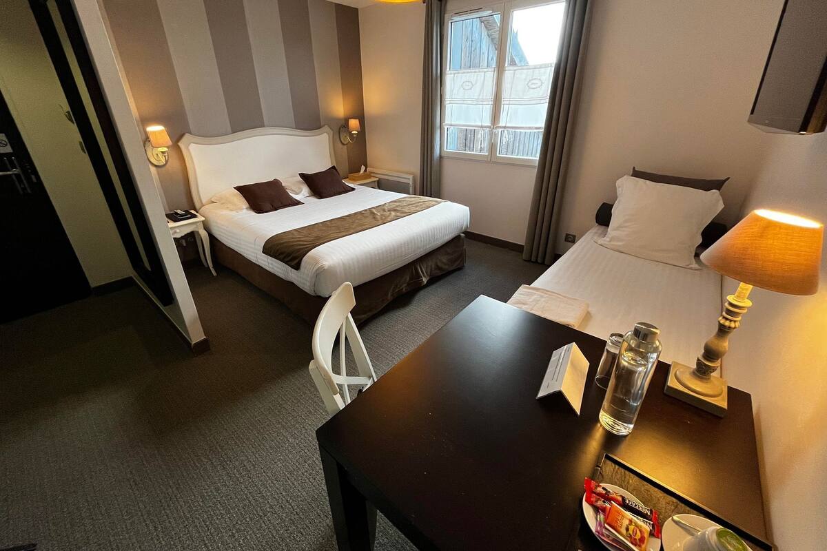 A hotel room with two beds, a desk, a chair, a window with curtains, and a lamp.
