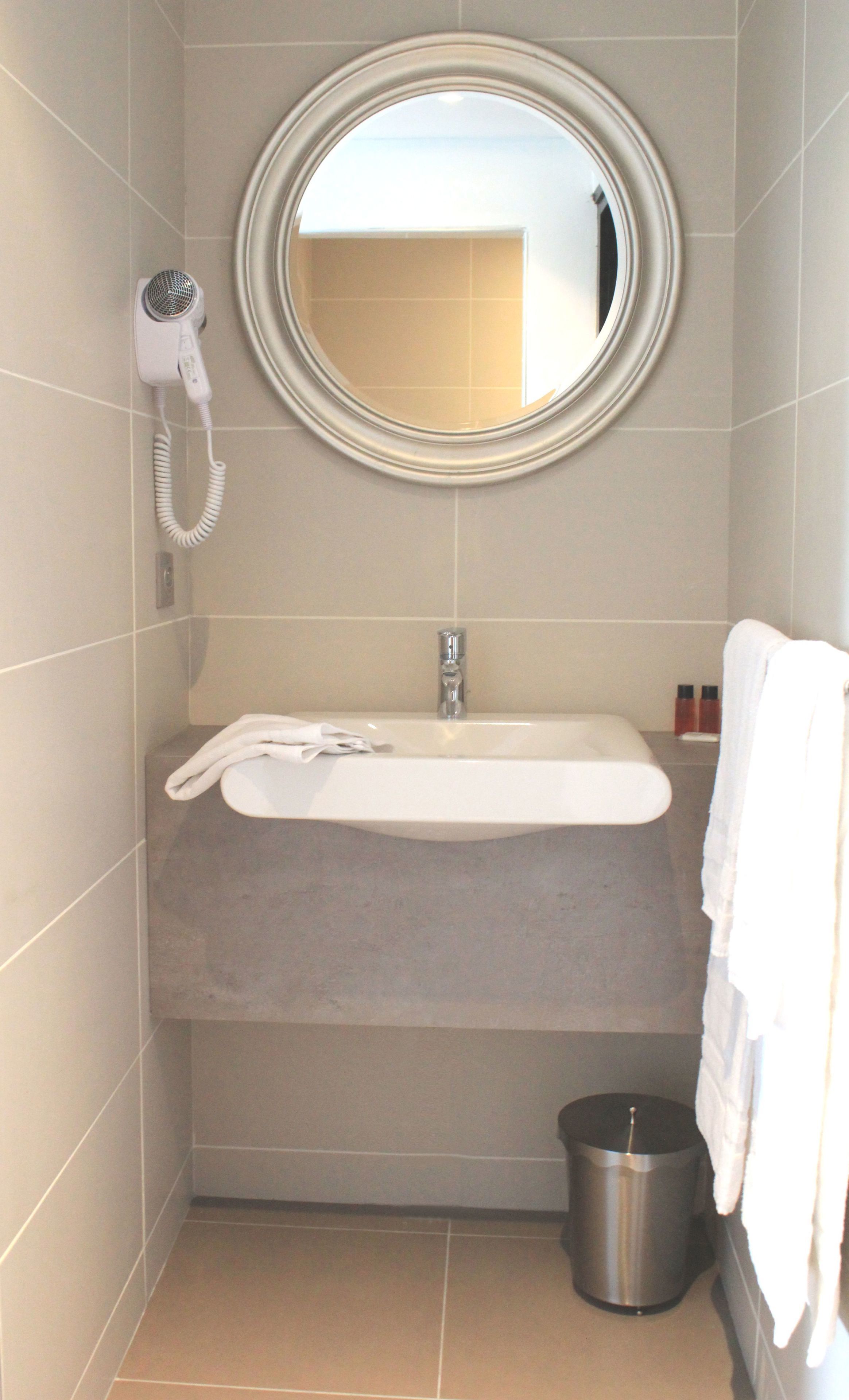 Une salle de bain équipée d’un miroir rond, d’un sèche-cheveux, d’un lavabo et d’une poubelle.