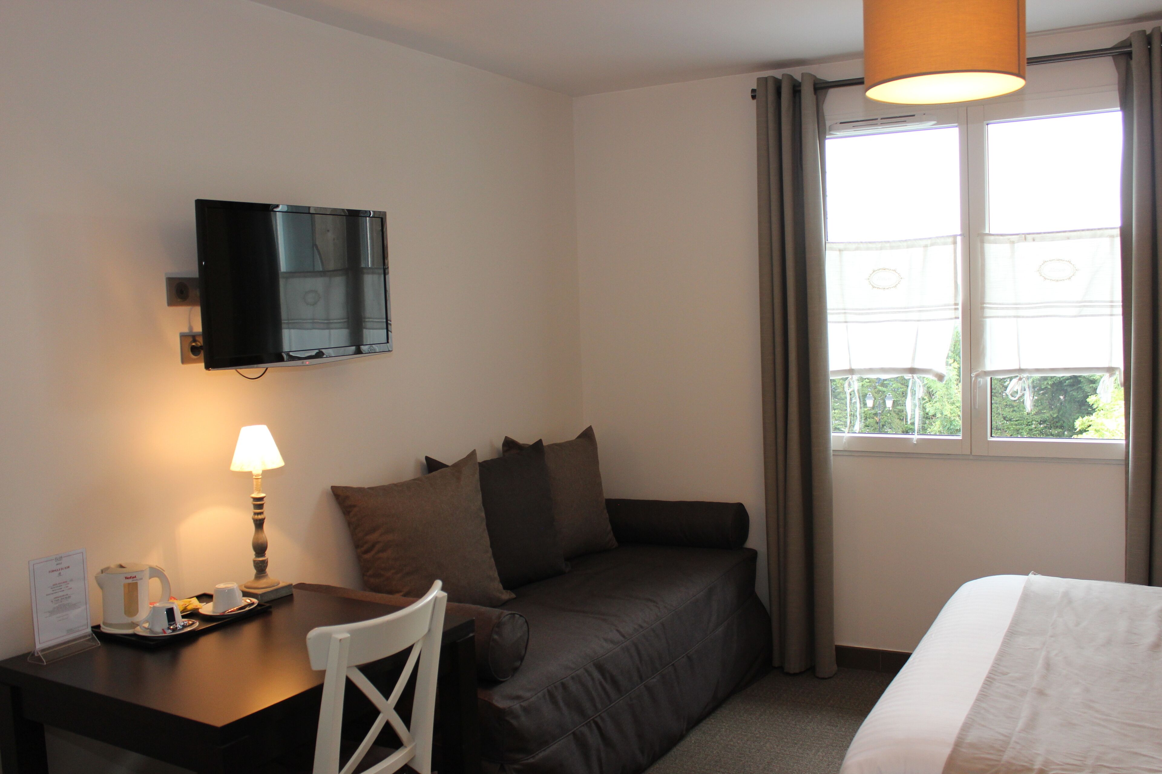 A hotel room with a bed, a sofa, a desk with a lamp, a television, and a window with curtains.