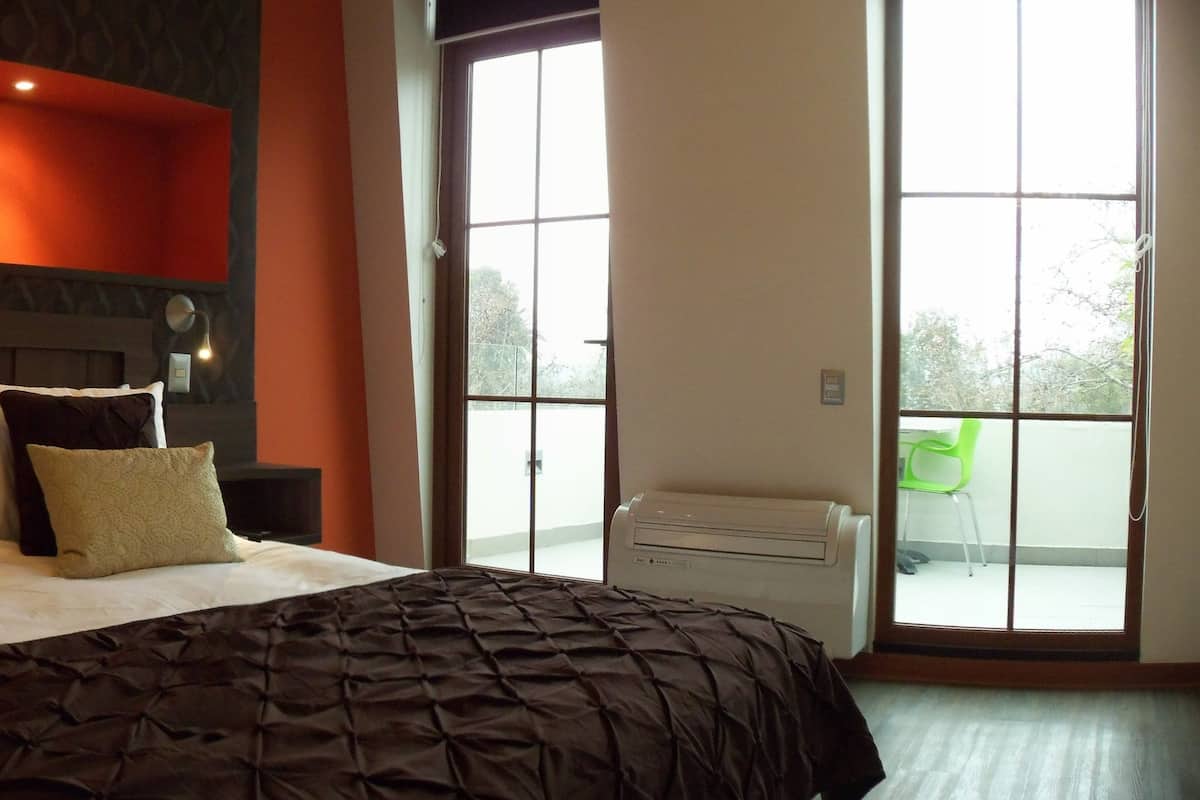 A bedroom with a large bed, a nightstand, a wall-mounted heater, and a window with a view of the outdoors.