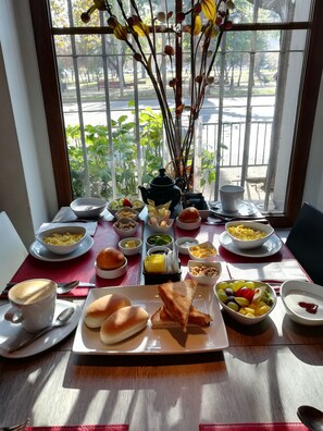 Una mesa de desayuno con diferentes platos y una tetera, frente a una ventana con vistas a una calle y follaje.