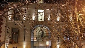 A building with a large arched window, a door, and a clock. There are people standing outside and a car parked in front. - SU Merced Boutique Hotel (SANTIAGO DE CHILE)