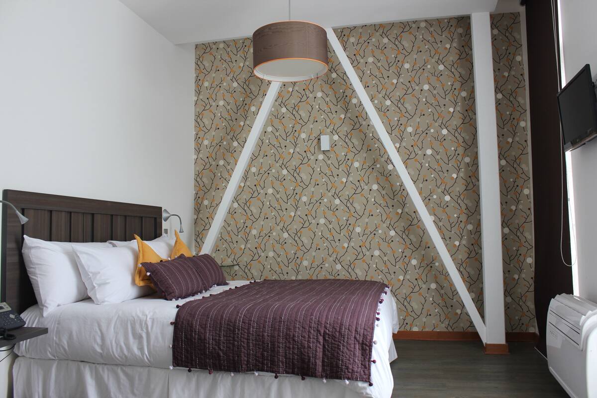 A bedroom with a bed, a bedside table, a lamp, a TV, and a patterned wallpaper.
