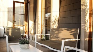 An outdoor seating area with white metal chairs and a table, a potted plant, and a building with large windows and a brick wall in the background. - SU Merced Boutique Hotel (SANTIAGO DE CHILE)