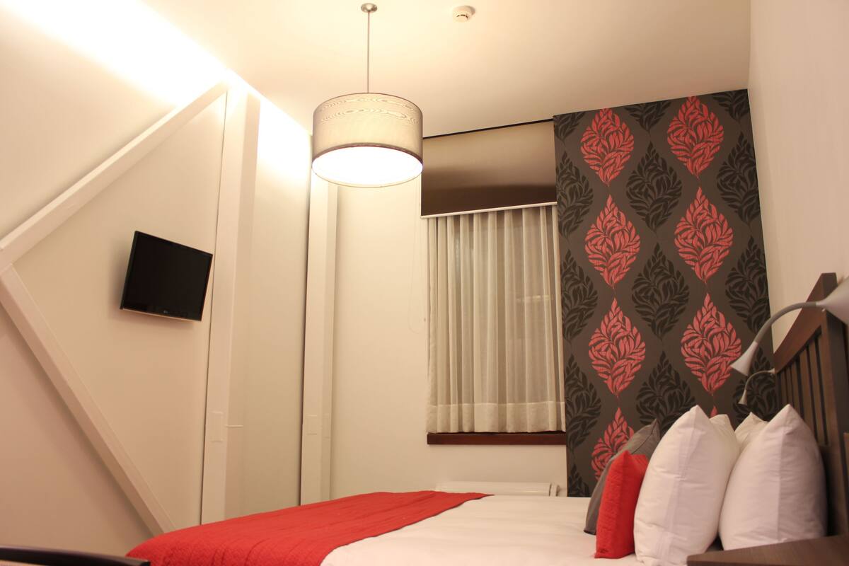 A hotel room with a bed, a television mounted on the wall, a window with curtains, and a patterned wallpaper.