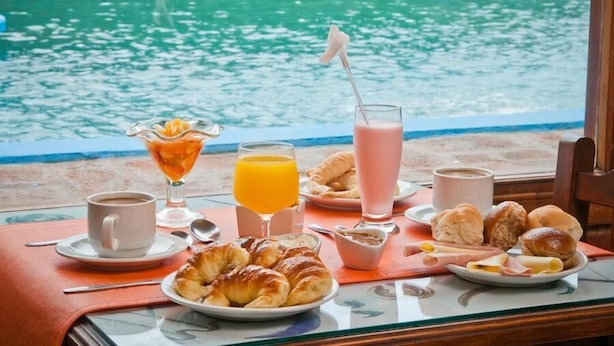 Une table de petit-déjeuner avec des croissants, des viennoiseries, du jus de fruits, du café et une boisson rose, le tout installé près d’une piscine.