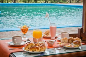 A breakfast table with croissants, pastries, juice, coffee, and a pink drink by a poolside. - Ciro (Pinamar)