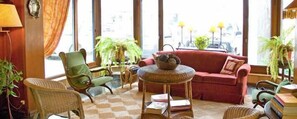 A cozy living room with a red sofa, green armchair, and wicker chairs. There are books on a small round table and potted plants around. - Hosteria Venezia (Pinamar)