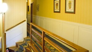 Una escalera con barandillas de madera y alfombra estampada, que lleva a una habitación con paredes amarillas y obras de arte enmarcadas.