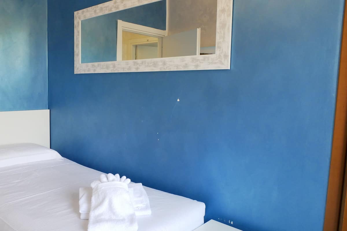 A single bed with white bedding and a white towel, a white-framed mirror on the wall, and a light switch on the wall.