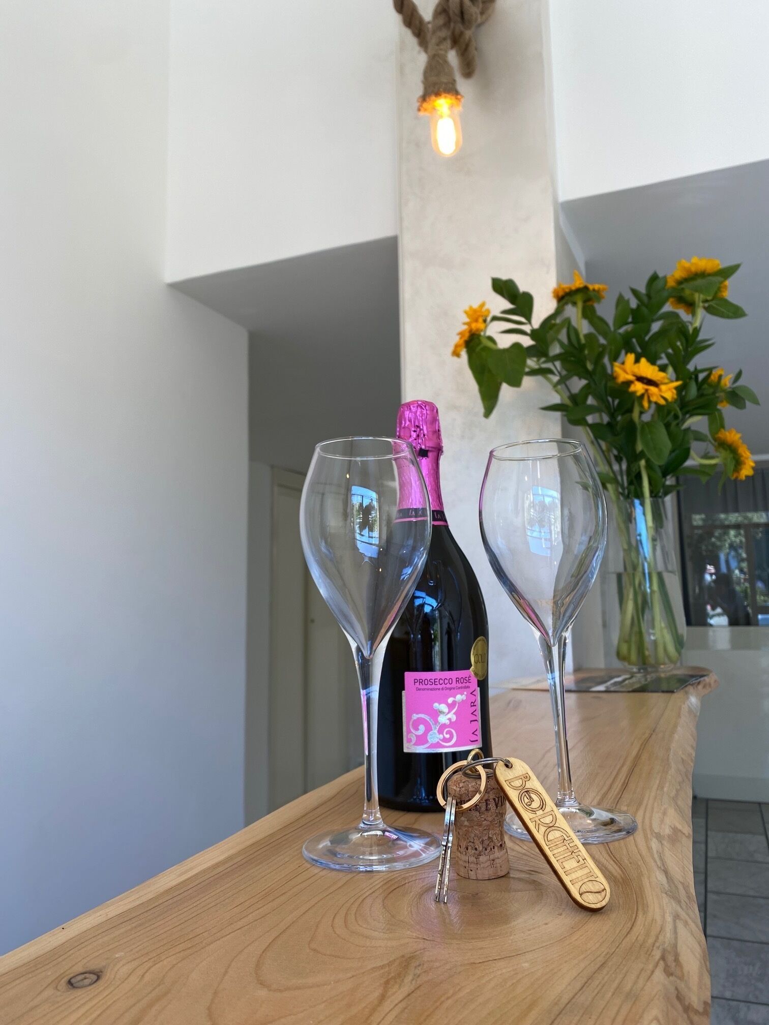 Ein Holztisch mit zwei Weingläsern, einem Korkenzieher und einer Flasche Prosecco Rosé in einem modernen Innenraum mit einer Betonstütze und einem Strauß gelber Blumen.