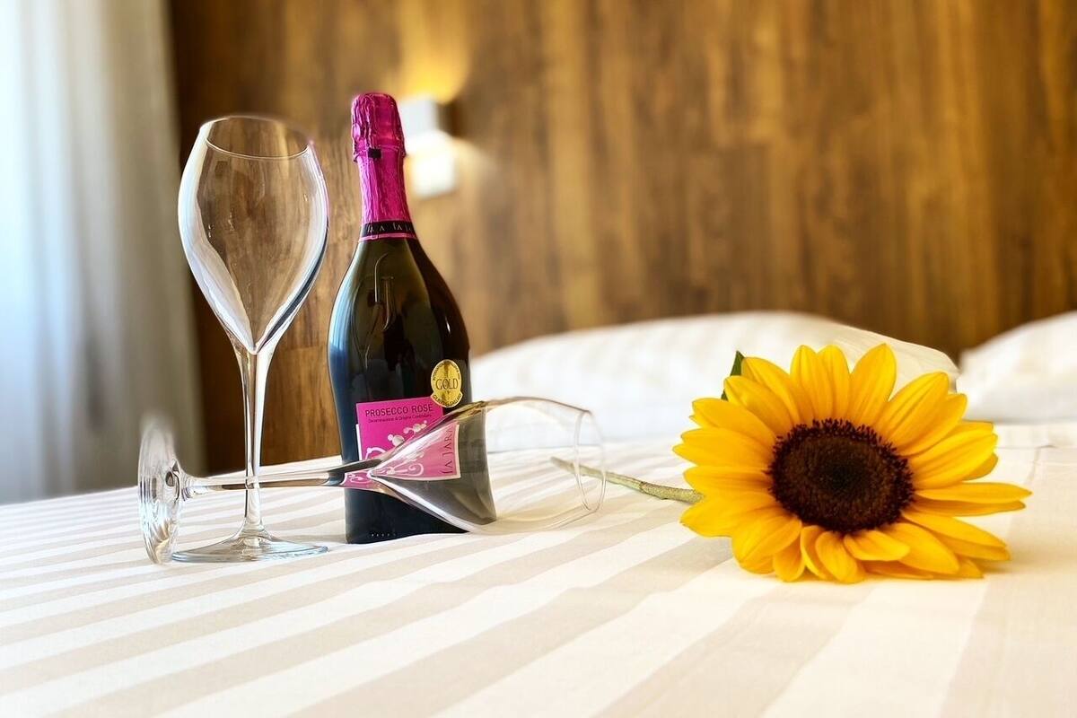 A bed with a striped bedspread, a bottle of Prosecco Rose wine, two wine glasses, and a sunflower.