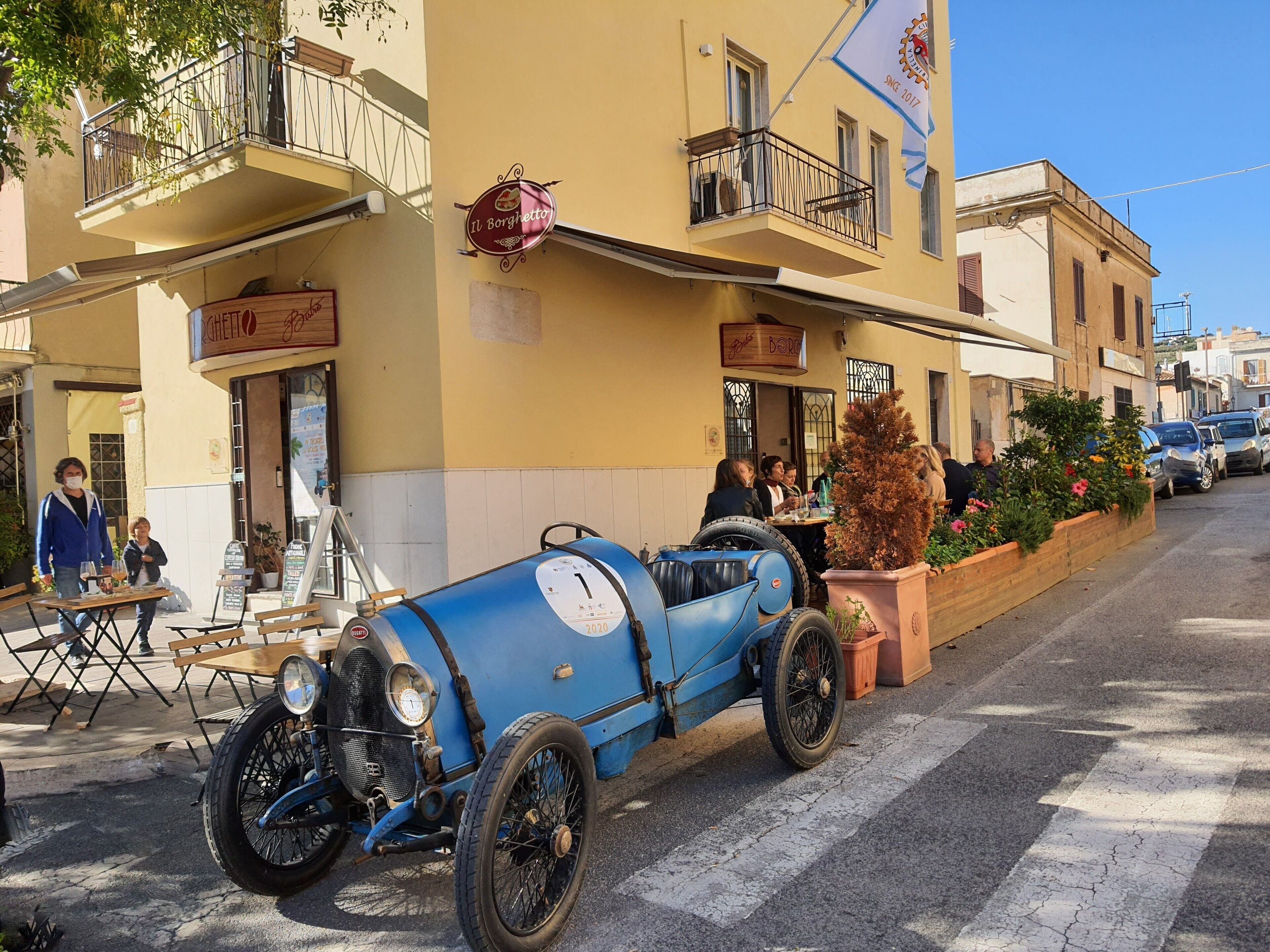 A vintage blue car parked in front of a yellow building with balconies and outdoor seating.