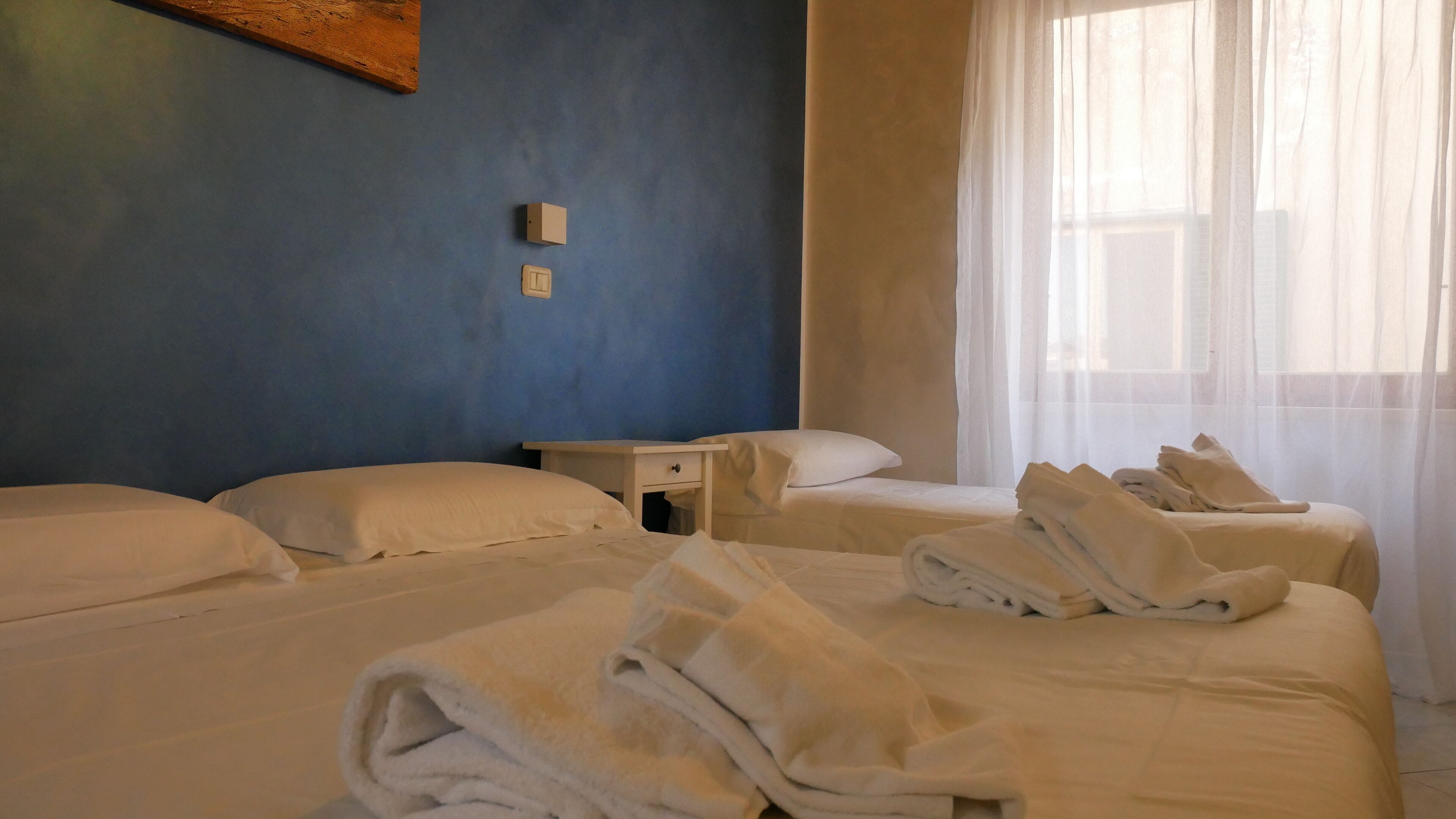Een hotelkamer met twee bedden, een nachtkastje en een raam met dunne gordijnen.