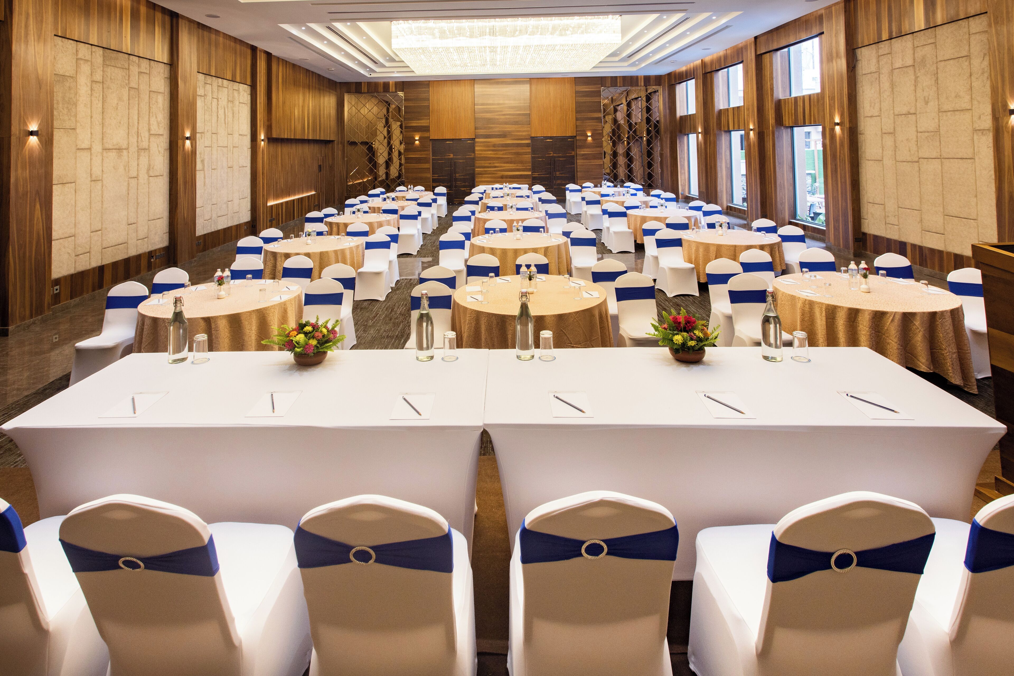 Un salón de conferencias con mesas redondas y sillas blancas, cada una adornada con cintas azules. Las mesas están preparadas con botellas de agua, vasos y blocs de notas.