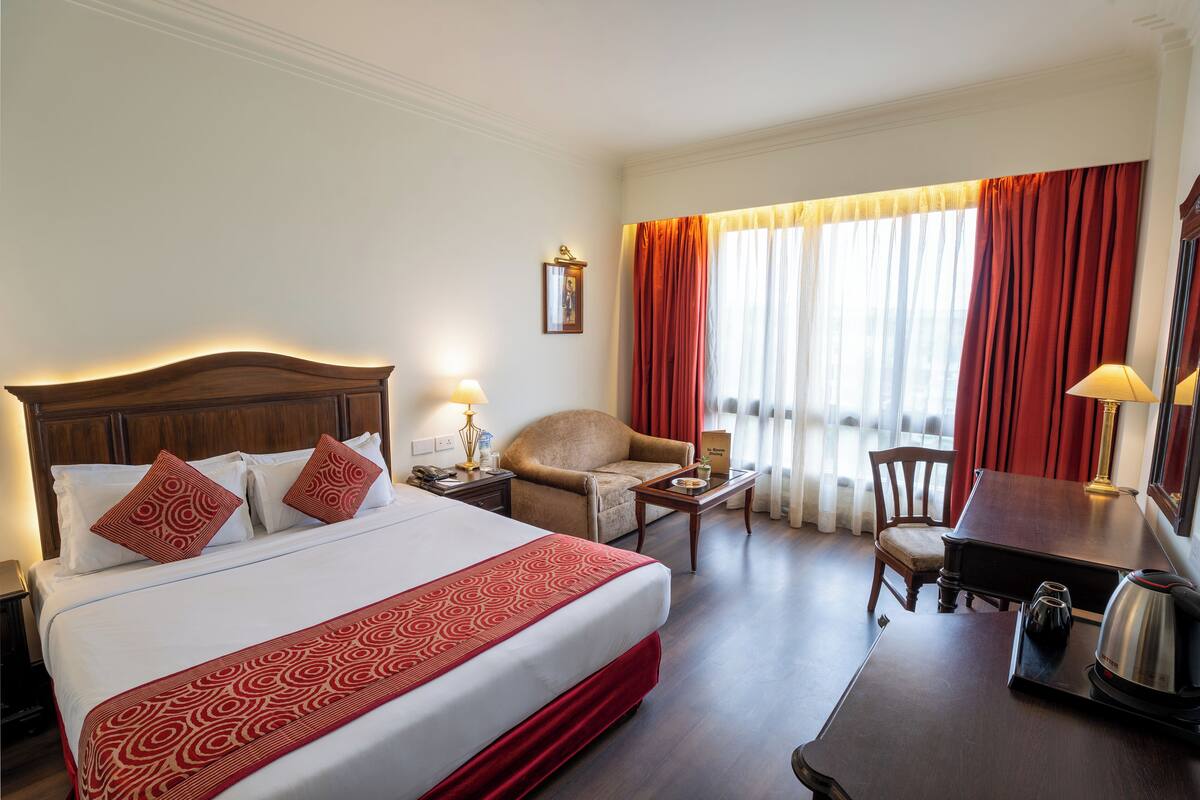 A hotel room with a large bed, a wooden headboard, a desk with a chair, a table with a lamp, and a window with curtains.