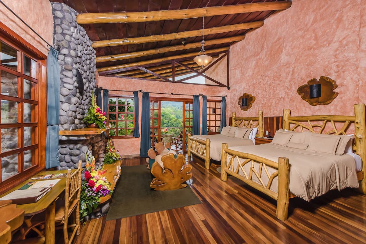 A rustic room with a stone wall, wooden ceiling beams, a bed, a dining table, and a view of greenery outside.