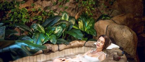 A woman relaxing in a hot tub surrounded by lush greenery and rocks.