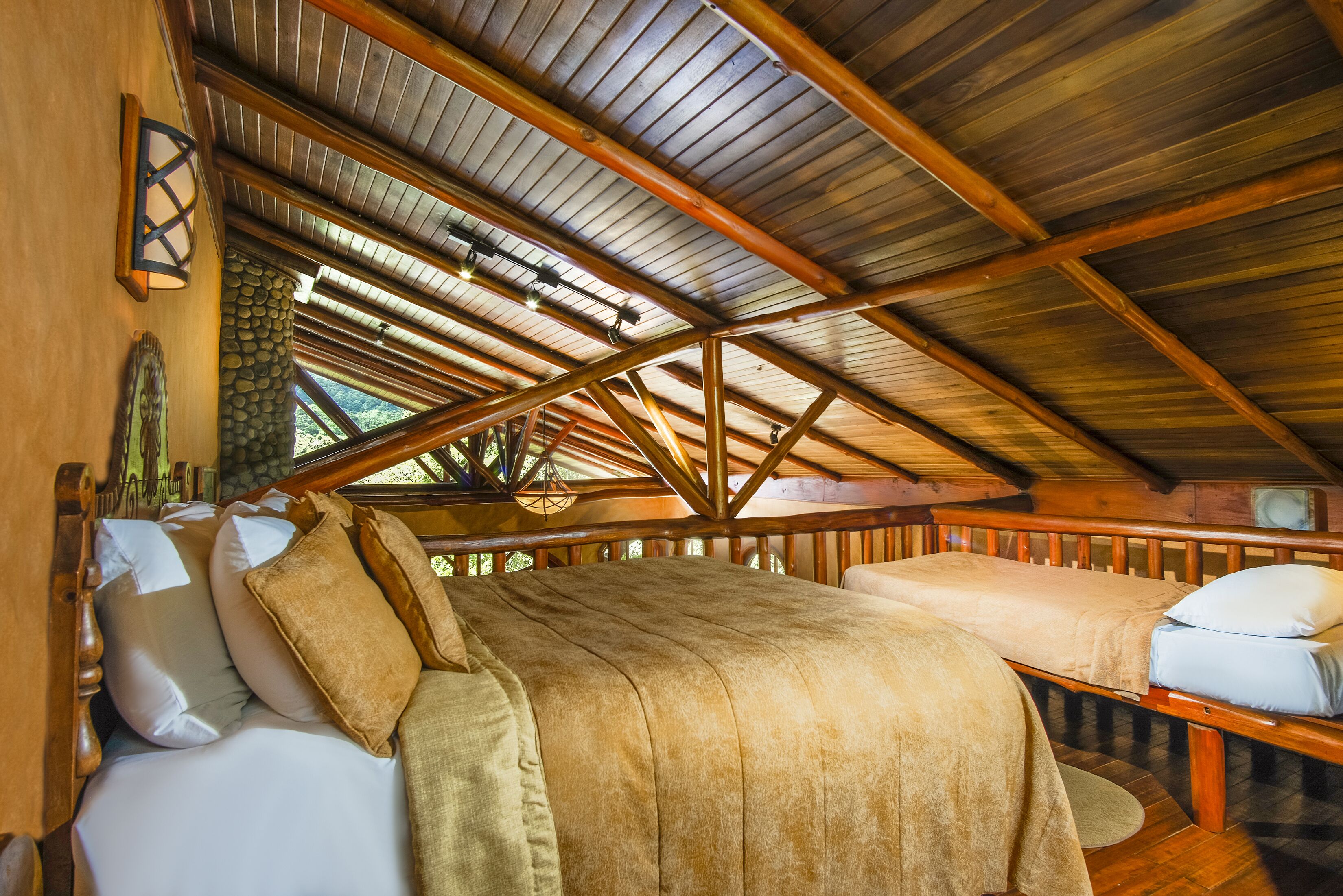 Une cabane en bois avec un plafond en pente, un lit recouvert d’une couverture beige et un mur en pierre.
