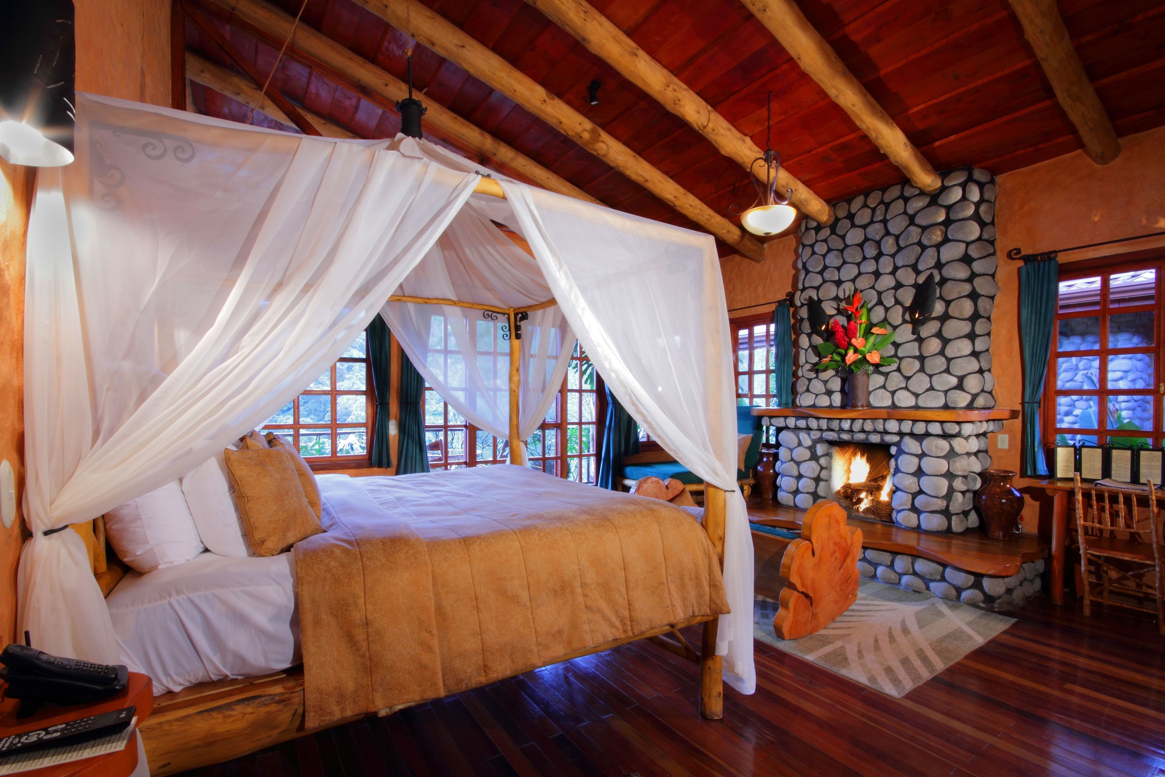 A spacious room with a large bed, a stone fireplace, and wooden beams on the ceiling.