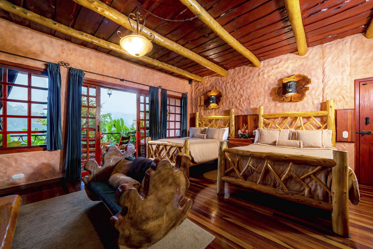 A rustic wooden room with a large bed, a wooden chair, and a view of greenery outside.