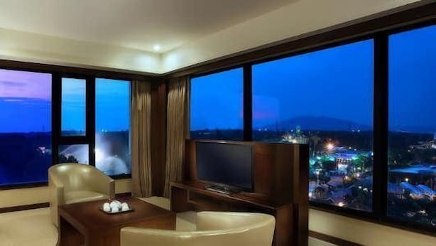 A hotel room with a large window offering a city view at dusk, a flat-screen TV, a desk with a lamp, and a sofa.