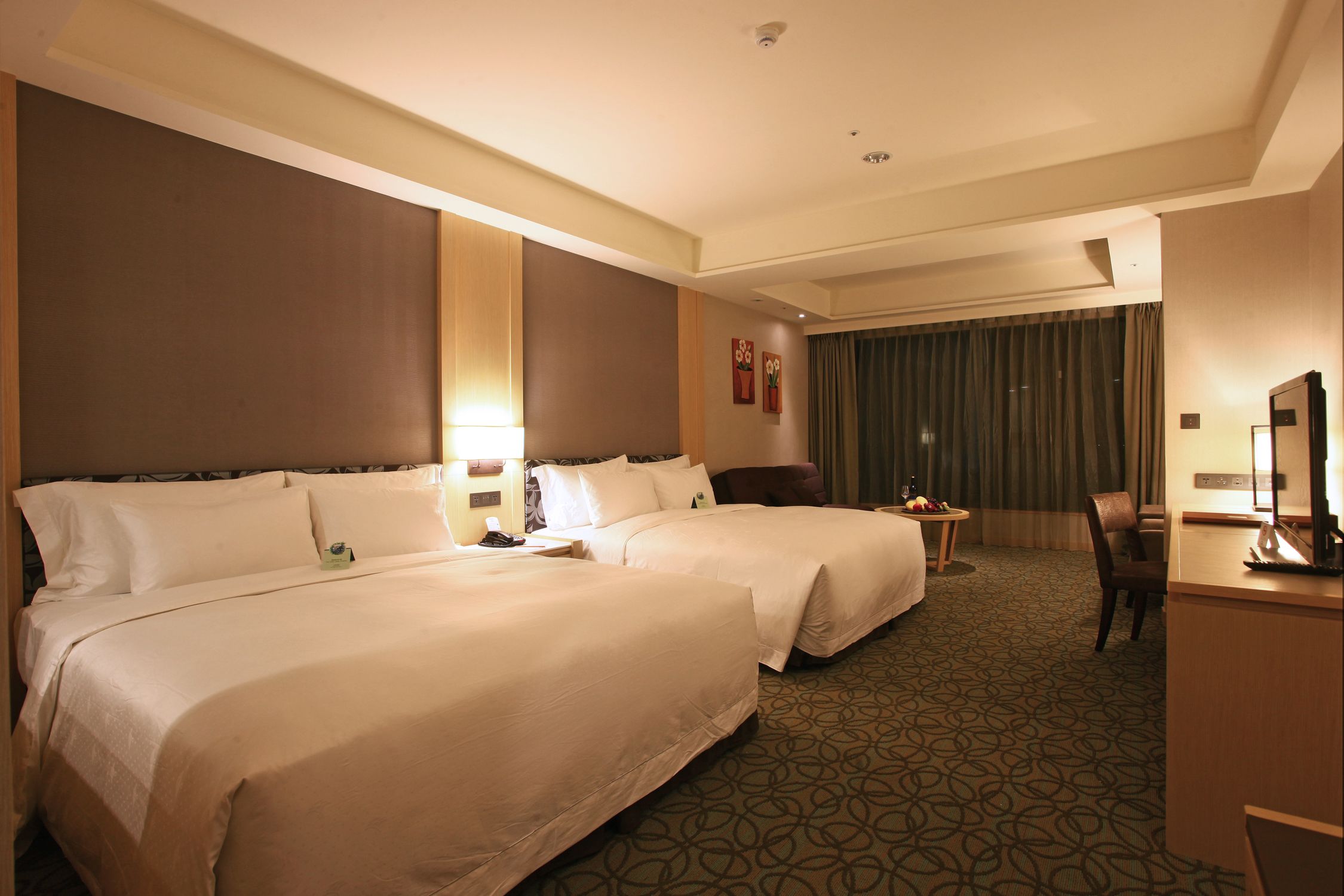 A hotel room with two beds, a desk, a chair, a small table with a fruit bowl, and a large window with curtains.