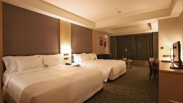 A hotel room with two beds, a desk, a chair, a small table with a fruit bowl, and a large window with curtains.