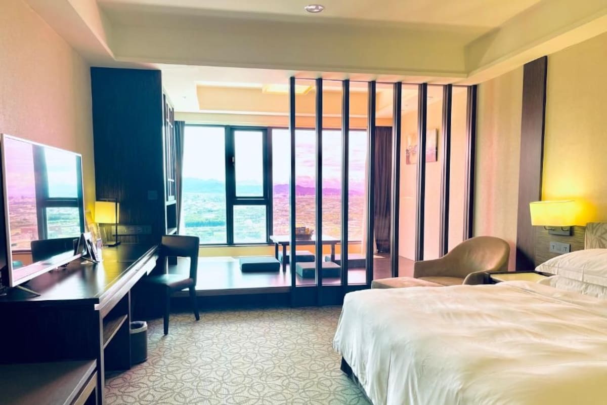 A hotel room with a bed, desk, chair, and a view of the outdoors.