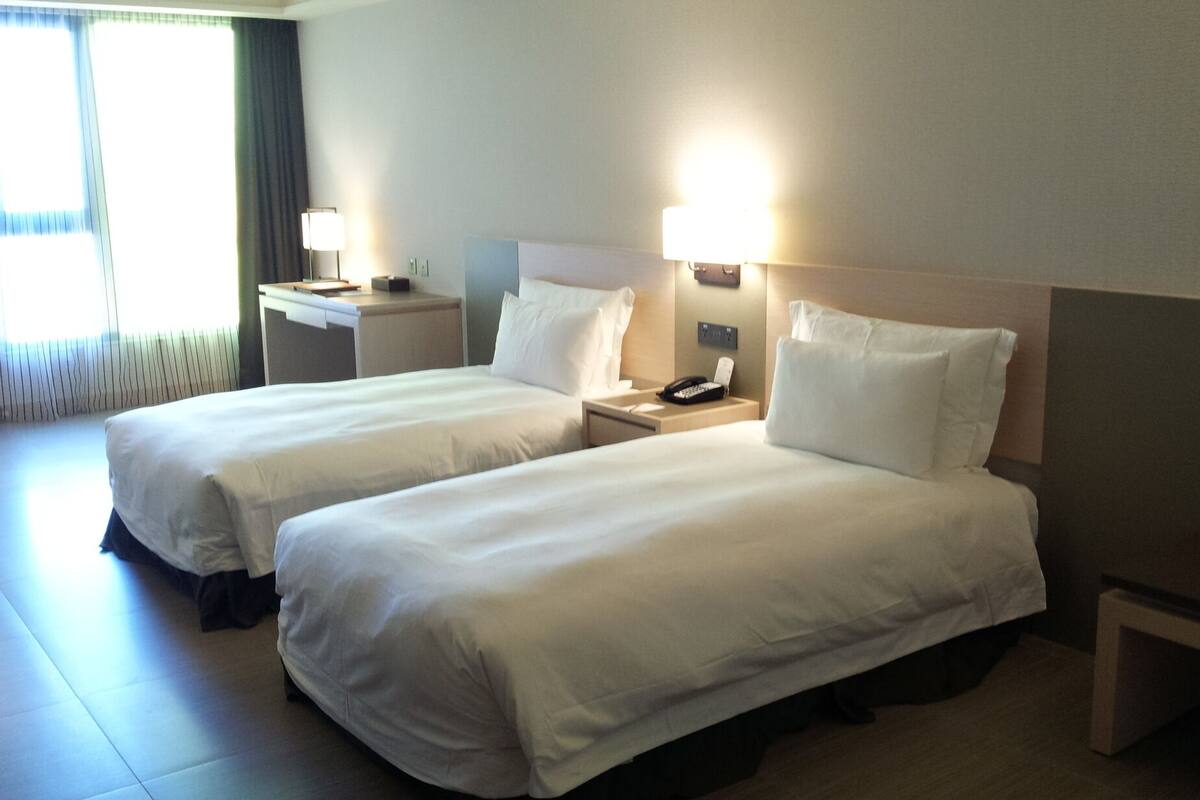 Ein Hotelzimmer mit zwei Betten, Nachttischlampen, einem Nachttisch und einem fensterbelaubten Fenster.