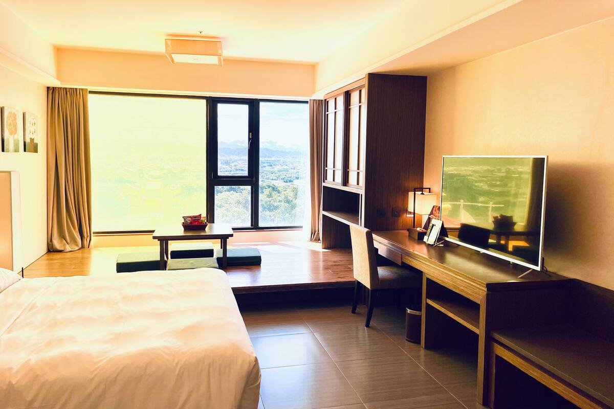 Een hotelkamer met een groot bed, een bureau met een televisie, een raam met uitzicht op de stad en een kleintje tafeltje met een zakje snacks.