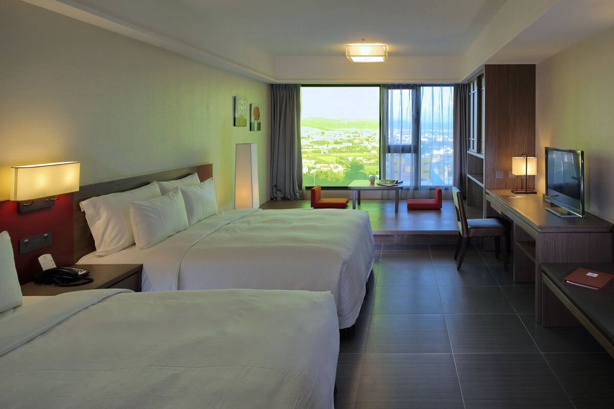 Ein Hotelzimmer mit zwei Betten, einem Schreibtisch, einem Fernseher und Blick auf eine Stadtlandschaft.