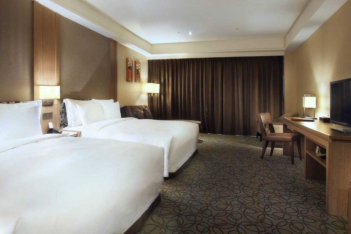 Ein Hotelzimmer mit zwei Betten, einem Schreibtisch, einem Fernseher und einem großen Fenster mit Vorhängen.