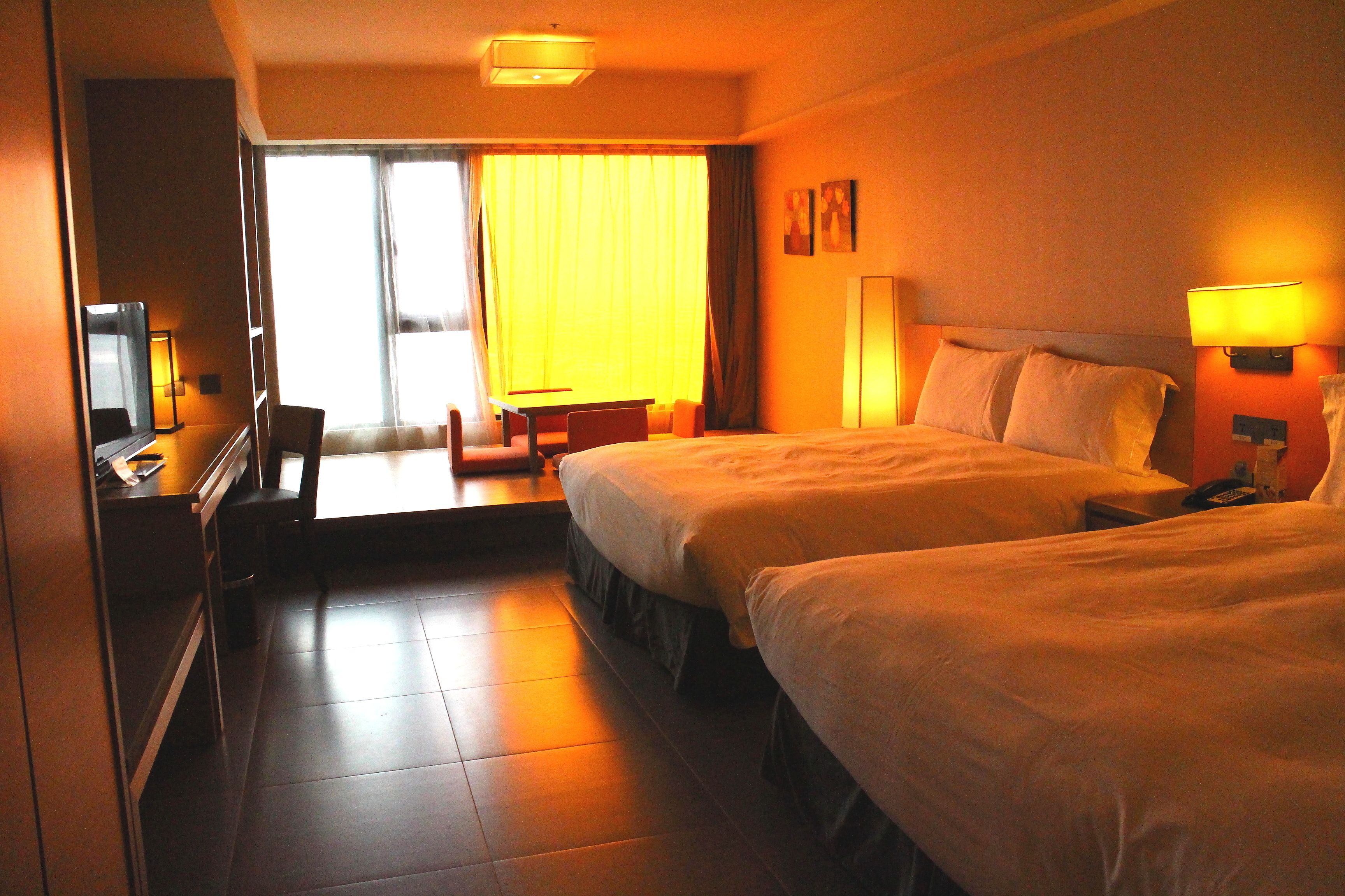A hotel room with two beds, a desk, a chair, and a view of the outdoors through a large window.