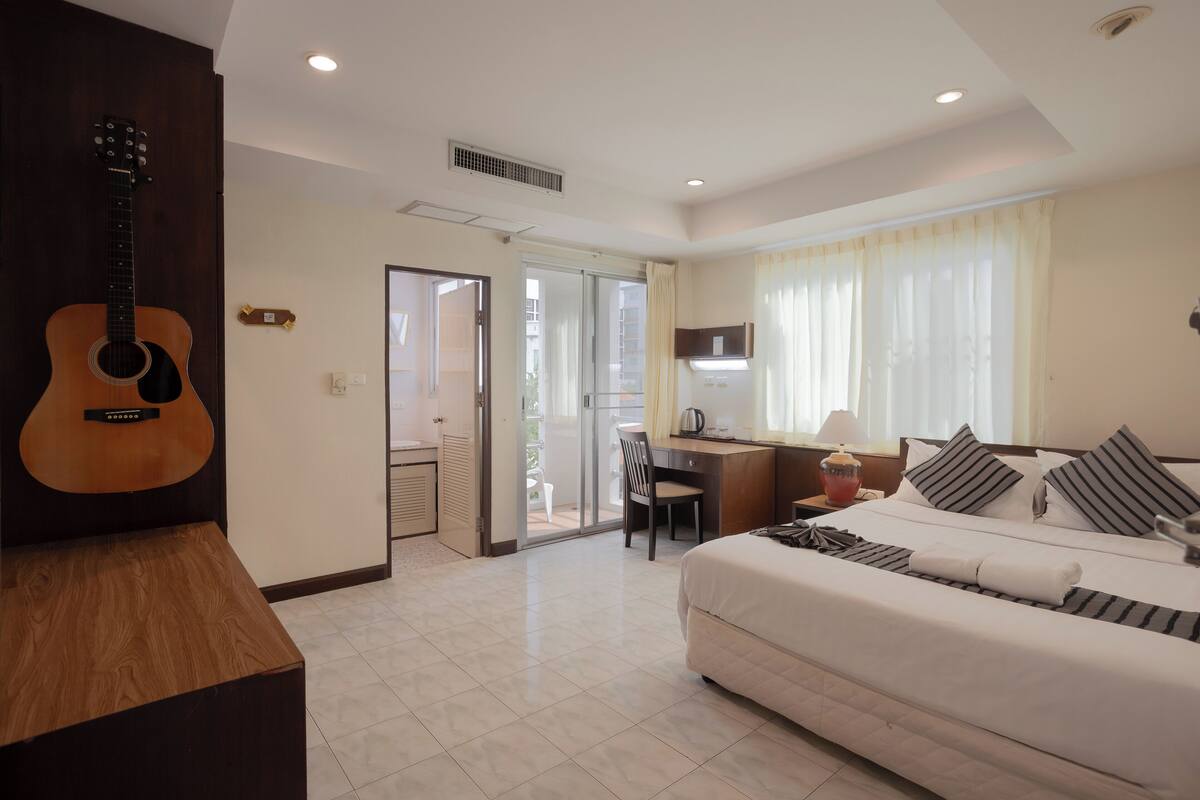 A hotel room with a bed, a desk, a guitar, and a sliding door to a balcony.