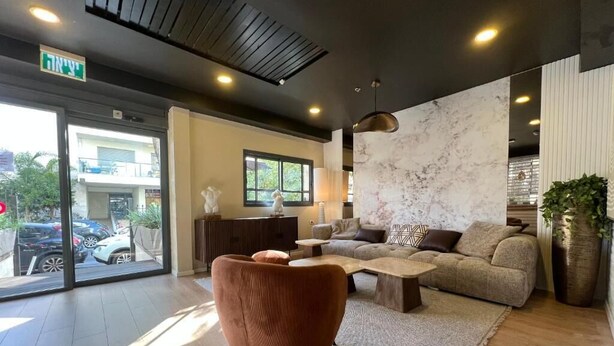 A modern living room with a sofa, armchair, and coffee table. There is a large window with a view of parked cars and greenery outside. - Beer Garden Hotel (Tel Aviv)